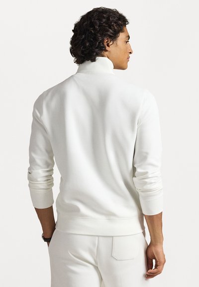 Uomo con capelli ricci che indossa un dolcevita bianco a maniche lunghe e pantaloni bianchi, visto di spalle.