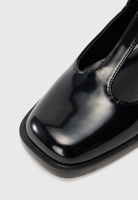 Pantofii din piele lacuită neagră cu vârf pătrat și detalii decupate, textură netedă și talpă subțire neagră.