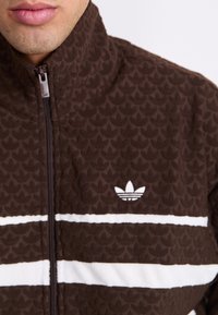 Καφέ ζακέτα με φερμουάρ, με μοτίβο υφής, που διαθέτει λευκές οριζόντιες ρίγες και ένα μικρό λευκό λογότυπο στην αριστερή πλευρά.