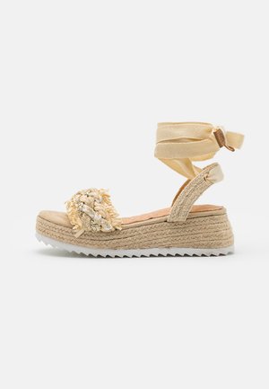 Beige espadrille wedgesandal med flettet frontrem, ankelbindinger og struktureret sål på en ensfarvet hvid baggrund.