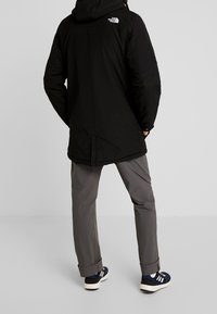 Personne portant une veste à capuche noire North Face, un pantalon gris et des baskets bleu marine avec des rayures blanches, debout de dos sur un fond uni.