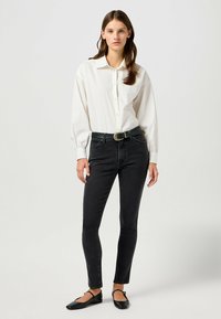 Camisa blanca de botones con mangas largas, jeans negros de tiro alto y bailarinas negras. El cinturón tiene un detalle decorativo en la hebilla de plata.