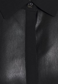 Chemise en faux cuir noir avec une surface texturée, un col classique et une fermeture à boutons. Le matériau est lisse avec un léger éclat.