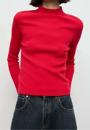 Mujer con suéter ajustado de cuello alto de manga larga rojo y vaqueros azul oscuro, de pie sobre un fondo claro y liso.