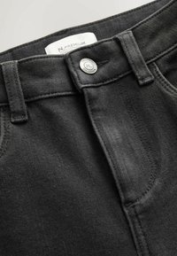 Czarne dżinsy z denimu na zbliżeniu, pokazujące pas, szlufki na pasek, guzik z napisem "N. Premium Collection" oraz częściowo widoczną białą wewnętrzną metkę.