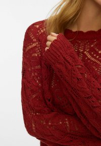 Maglione rosso lavorato a maglia con un motivo di pizzo, polsini a costine e scollatura a onda. La texture è leggera e a maglia aperta.