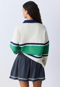 Mujer con cabello largo y rizado, vestida con un suéter de rayas blancas, verdes y azul marino y una falda tableada azul marino, vista desde atrás.