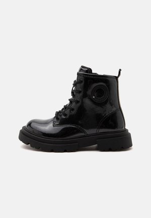 Black shiny lace-up combat boot with thick sole and circular patch detail on the side, shown from the outer side on a white background.