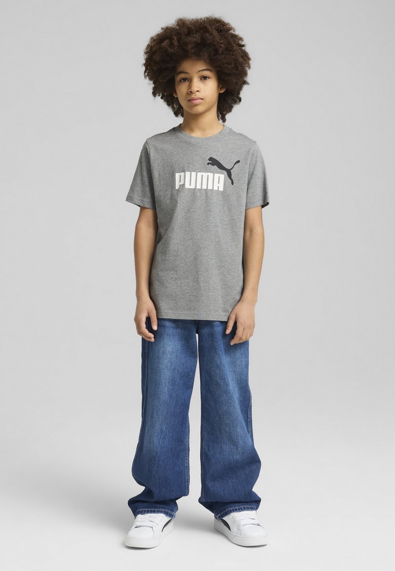Pelēks kokvilnas T-krekls ar baltu "PUMA" logotipu, apvienots ar brīvi pieguļošu zilu džinsu biksēm un baltām kedām. Stāv uz neitrāla fona.
