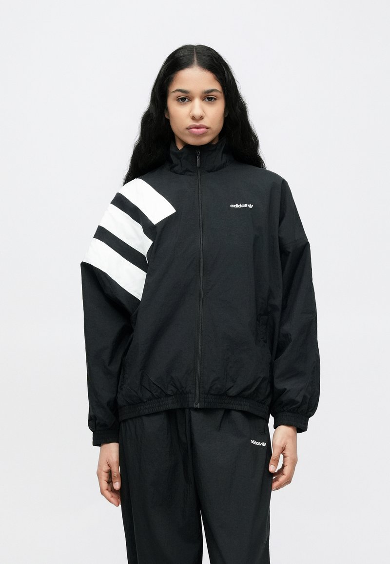 Sort vindjakke med høj krave, der har et hvidt diagonalt stribemønster på venstre ærme og en broderet logo på brystet.