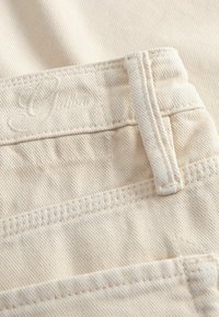Μπεζ denim ύφασμα με εμφανείς ραβδώσεις και ραφές. Διαθέτει ένα εμφανές λογότυπο ραμμένο κοντά στη μέση και έχει δομημένο σχήμα.