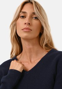 Granatowy sweter dzianinowy z dekoltem w serek. Miękka tekstura i dopasowane rękawy. Modelka dotyka dekoltu, ukazując detale odzieży.