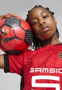 Raudonas futbolo kamuolys su juodais akcentais ir Puma logotipu, laikomas priešais tekstūruotą raudoną marškinėlį su juodomis detalėmis ir baltu užrašu.