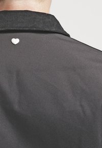 Camicia nera con colletto texture e una piccola spilla a forma di cuore bianco attaccata vicino al colletto sul retro.