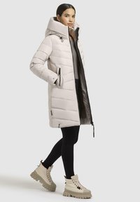 Light grey puffer coat with a high collar, zippered pockets, and ribbed accents. Paired with black leggings and beige chunky boots.