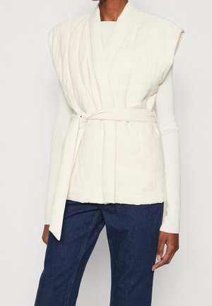 Persoon draagt een wit doorgestikt mouwloos vest met riem over een wit geribd shirt met lange mouwen en donkerblauwe spijkerbroek.