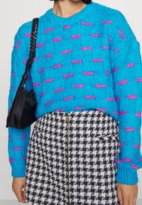 Modrý pletený svetr s růžovými stehy, sukně v černobílém houndstooth vzoru s předním zipem a černá kabelka přes rameno s pleteným popruhem.