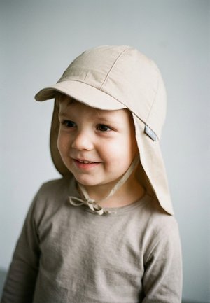 Kleines Kind lächelt, trägt einen beigen Sonnenhut mit Nackenschutz und Bindebändern, bekleidet mit einem beigen Langarmshirt, vor einem schlichten hellen Hintergrund.