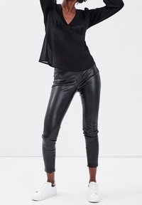 Henkilö, joka käyttää mustaa läpinäkyvää pitkähihasta ykköstä, mustia nahkahousuja ja valkoisia lenkkareita, seisoo kädet nostettuna valkoista taustaa vasten.