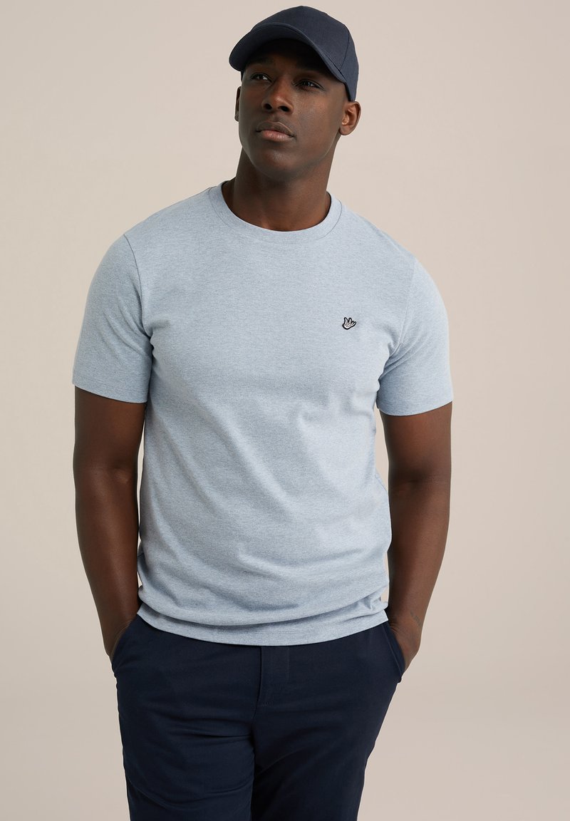 Gaiši zils īsās piedurknes T-krekls kokvilnas maisījumā, ar mazu melnu logotipu uz krūtīm. Modelis valkā tumši zilu cepuri un tumši zilus bikses.