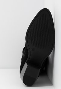 Bottine en daim noir avec une semelle en caoutchouc texturée, un petit talon bloc et une fermeture éclair latérale. L'embout est arrondi, et le design est simple sans motifs.