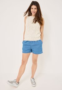 Top senza maniche di maglia color beige chiaro abbinato a pantaloni corti in denim blu. Il modello indossa sneaker grigie con accenti bianchi. Completo semplice e casual.