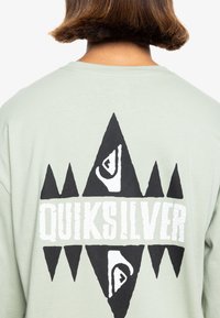 Light green cotton long-sleeve shirt featuring a large black and white graphic design with triangular shapes and the text "QUIKSILVER."