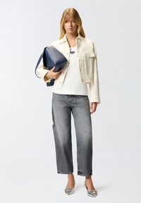 Top bianco a canotta sotto una giacca cropped color crema, jeans grigi a gamba larga e ballerine argento con punta affilata, mentre tiene una borsa blu navy.