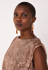 Top in paillettes rosate con scollo rotondo e maniche corte a cappuccio. Grandi orecchini con perle ambra e trasparenti offrono un contrasto. Texture liscia.