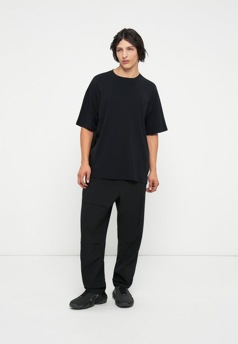 Camiseta negra de gran tamaño con cuello redondo, combinada con pantalones cargo negros que tienen múltiples bolsillos y calzado negro grueso.