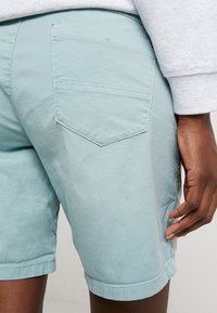 Personne portant un short bleu clair avec une poche arrière, la main reposant près de la cuisse droite, vêtue d'un haut à manches longues gris clair.
