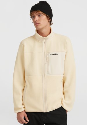 Young man wearing cream fleece jacket with zip-up front and chest pocket, black beanie, and black pants, looking to the side.