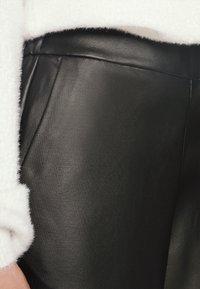 Närbild av svarta läderbyxor som bärs med en vit fluffig tröja, vilket framhäver midjebandet och sidsömmens detaljer.