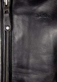 Gros plan sur une veste en cuir noir avec le logo Schott NYC en relief, une fermeture éclair en métal et un bouton-pression en métal marqué.