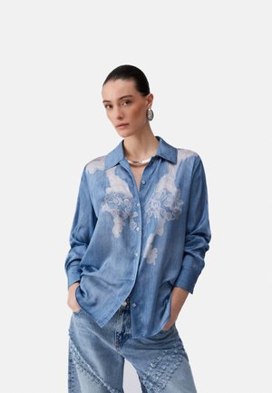 Mujer con el cabello peinado hacia atrás que lleva una camisa de mezclilla azul con bordado floral de encaje blanco y jeans de mezclilla a juego con parches, con las manos en los bolsillos.