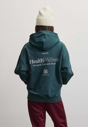Persona che indossa una felpa con cappuccio verde scuro con il testo "Health Wellness" sulla schiena e un cappello bianco lavorato a maglia, in piedi davanti a uno sfondo chiaro e uniforme.