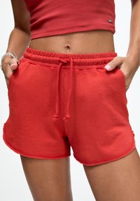Vrouw in rode drawstring shorts en een rode crop top met handen in de zakken, die haar middenrif en armbanden om haar linkerschouder toont.