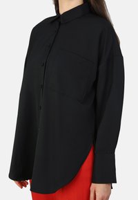Camisa de botones negra en un tejido ligero, con un bolsillo en el pecho, mangas largas y un dobladillo curvado. Diseño limpio y clásico.