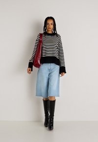 Sweater listrado em preto e branco com mangas longas, culotes de denim azul claro e botas pretas até ao joelho; a segurar uma bolsa vermelha brilhante.