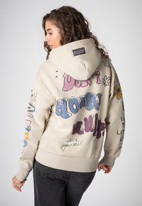 Beige hoodie gemaakt van zachte stof, met kleurrijke tekst en graphics, een grote capuchon, geribbelde manchetten en een relaxte pasvorm.