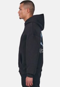 Black hooded sweatshirt with a loose fit, front pocket, and graphic accents on the back. Fabric appears soft and textured.