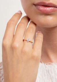 Hand with gold ring featuring four small diamonds in a flower shape, touching chin near lips, on light background.
