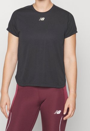 Kvinde iført en sort New Balance T-shirt med korte ærmer og bordeauxfarvede New Balance leggings stående imod en ensfarvet baggrund.