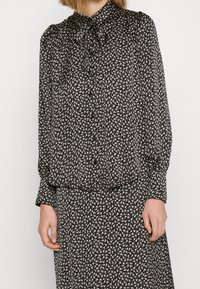 Blusa negra con flores con un cuello alto y lazo, que presenta pequeños patrones de flores color crema, y mangas largas con puños fruncidos.