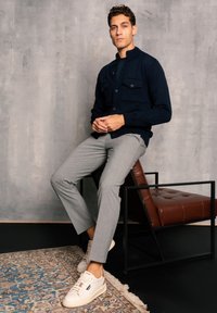 Navy knit cardigan with button closure and two front patch pockets, paired with light gray tailored pants and white sneakers on a patterned rug.