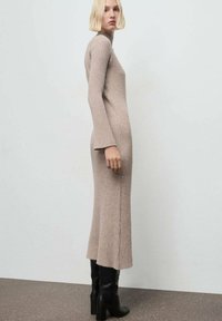 Abito lungo a maglia beige, rigato, con collo alto e maniche a campana, caratterizzato da fessure laterali, abbinato a stivali neri con tacco al ginocchio.
