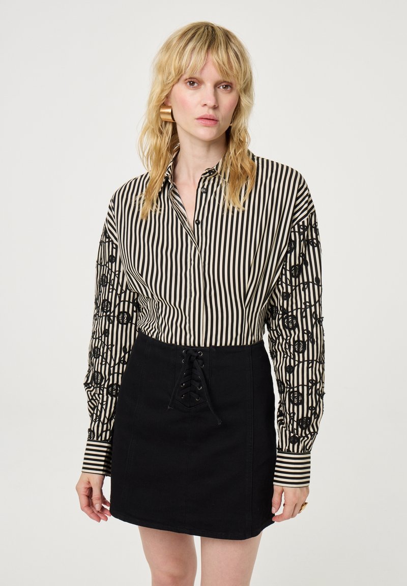 Gestreept zwart en beige shirt met knoopsluiting en zwarte patroonsmouwen, gecombineerd met een zwarte rok met een vetersluiting aan de voorkant.