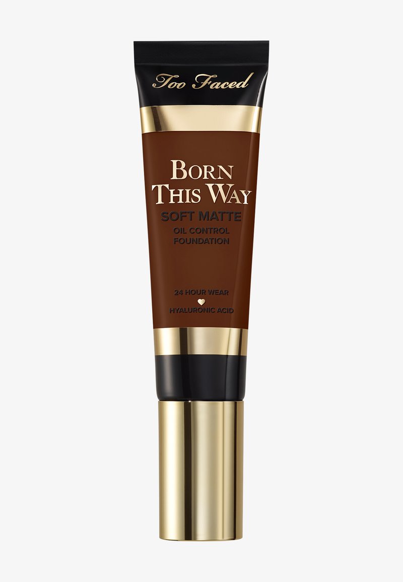 Tube de fond de teint avec un corps marron, des accents dorés et un capuchon noir. Texte : "Fond de teint mat à contrôle de l'huile Born This Way." Tenue de 24 heures.