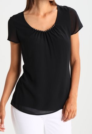 Femme portant un chemisier noir à manches courtes avec un détail de perles au col et un pantalon blanc, debout devant un fond clair uni.