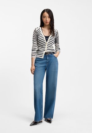 Vrouw draagt een zwart-wit gestreepte cardigan, blauwe jeans met hoge taille en wijde pijpen, een zwarte riem en zwarte puntige schoenen, staand met één hand in de zak.